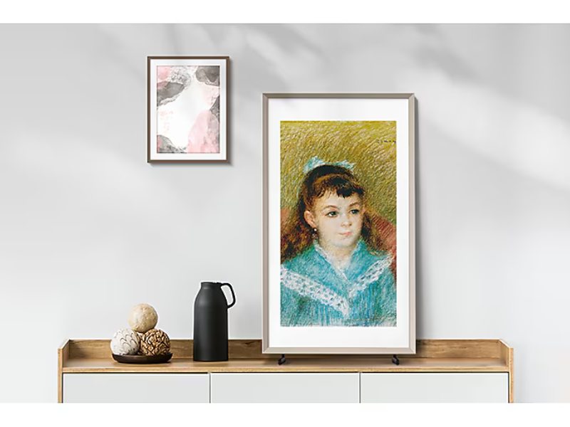 Samsung The Frame 32-inch Full HD QLED TV displaying digital art, placed on a wooden sideboard next to a black jug and decorative balls; above, a small abstract artwork hangs on a light gray wall.