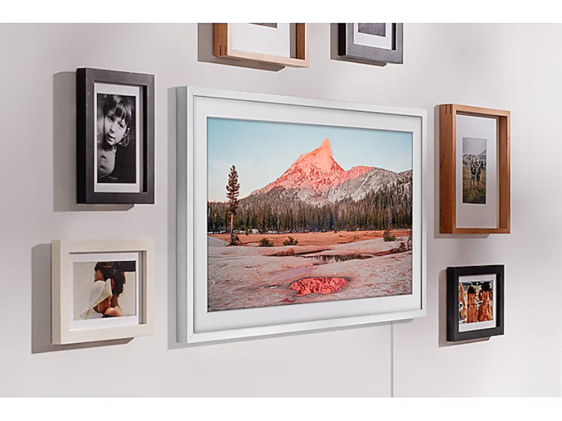 Living room wall featuring a 32-inch Samsung The Frame QLED-TV displaying a pink sky mountain landscape, centrally placed among variously colored framed portrait and landscape photos. Modern gallery-style TV blends seamlessly with decorative photo frames for stylish home décor.