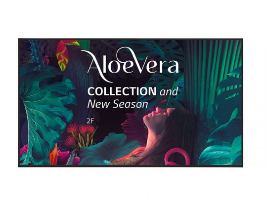 Promotionsposter für die Aloe Vera COLLECTION New Season - eine Frau sitzt umgeben von üppigen, tropischen Pflanzen und bunten Blättern in leuchtendem Grün, Blau und Pink. Auf dem Poster steht außerdem "2F". Ideal als Werbebild für natürliche Hautpflege oder Modekollektion mit Aloe Vera.