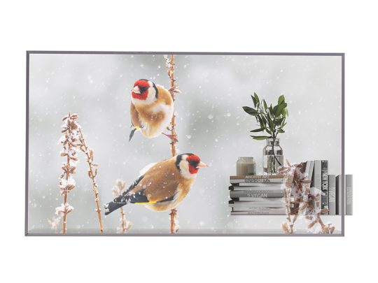 Zwei bunte Stieglitze sitzen auf trockenen Stängeln im Schneefall. Im Vordergrund stehen mehrere Bücher, eine kleine Vase mit grünen Blättern und dekorative Gegenstände auf einem Regal - eine harmonische Kombination aus Naturmotiv und stilvoller Wohnungsdekoration.
